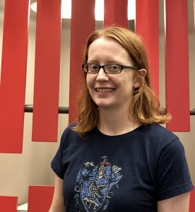 A red-haired white woman with glasses in front of a geometric background