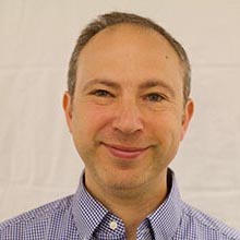 A clean-shaven man with fair skin and short gray hair wearing a blue button-down shirt