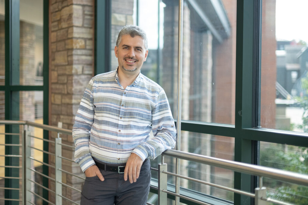 A smiling man with graying hair and a short beard, wearing a striped shirt, stands at a railing in front of a large window.