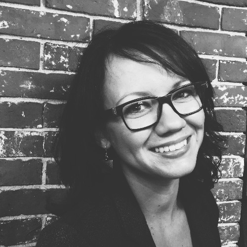 Black-and-white photo of a smiling woman with fair skin and dark hair, wearing glasses, in front of a brick wall