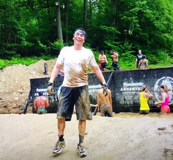 A man with dark hair wearing very muddy athletic clothes smiles in front of a muddy pond full of other racers and a black wall