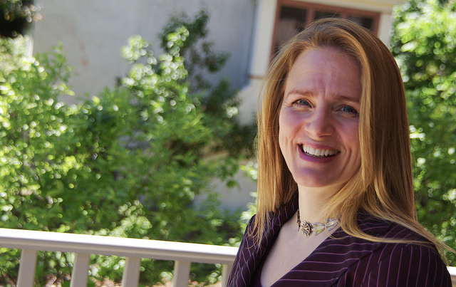 A white woman with long blond hair wearing a striped maroon jacket poses on a deck near trees