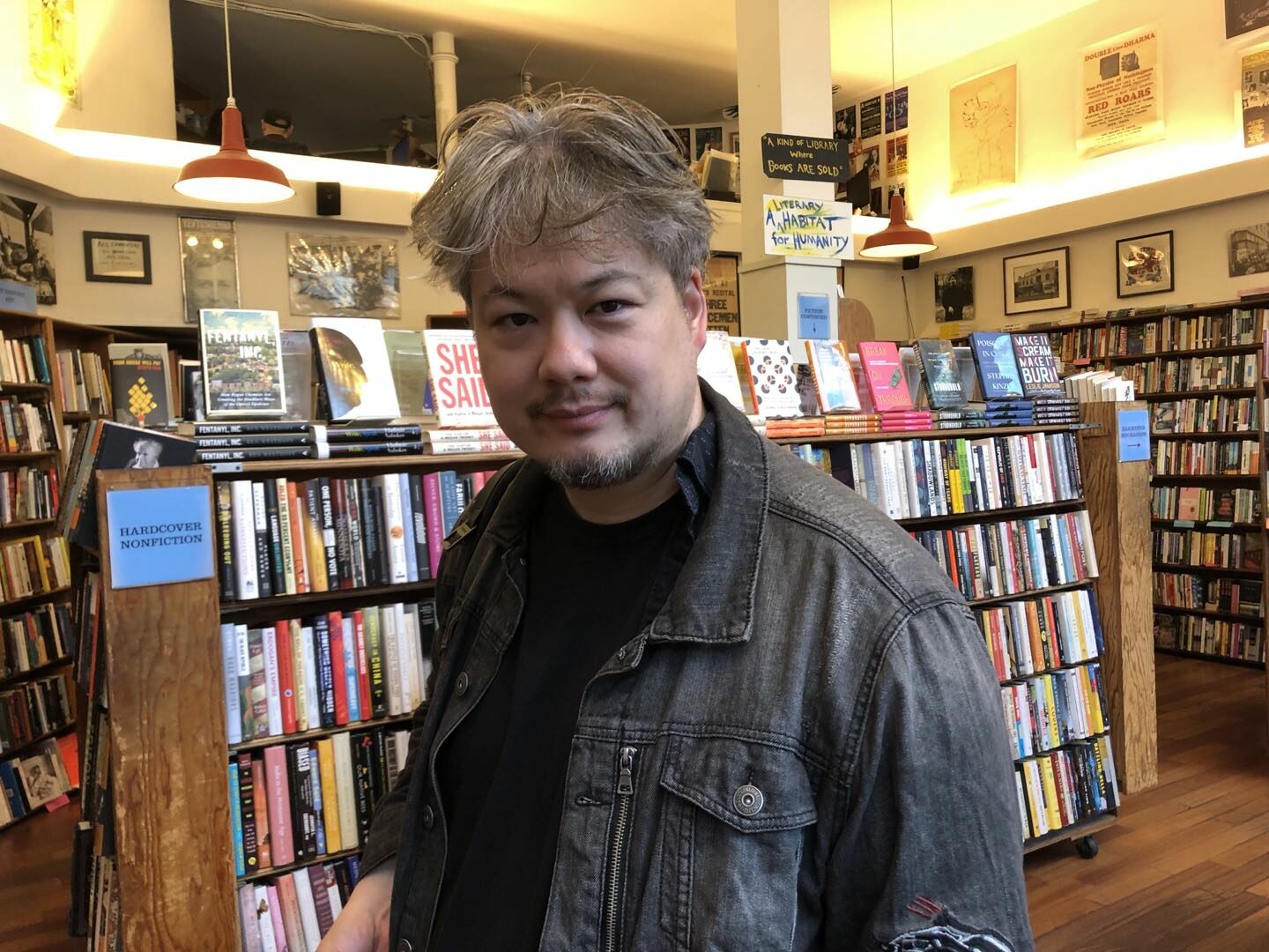 A man with dark graying hair and a small goatee beard standing in front of a full bookshelf in a bookstore