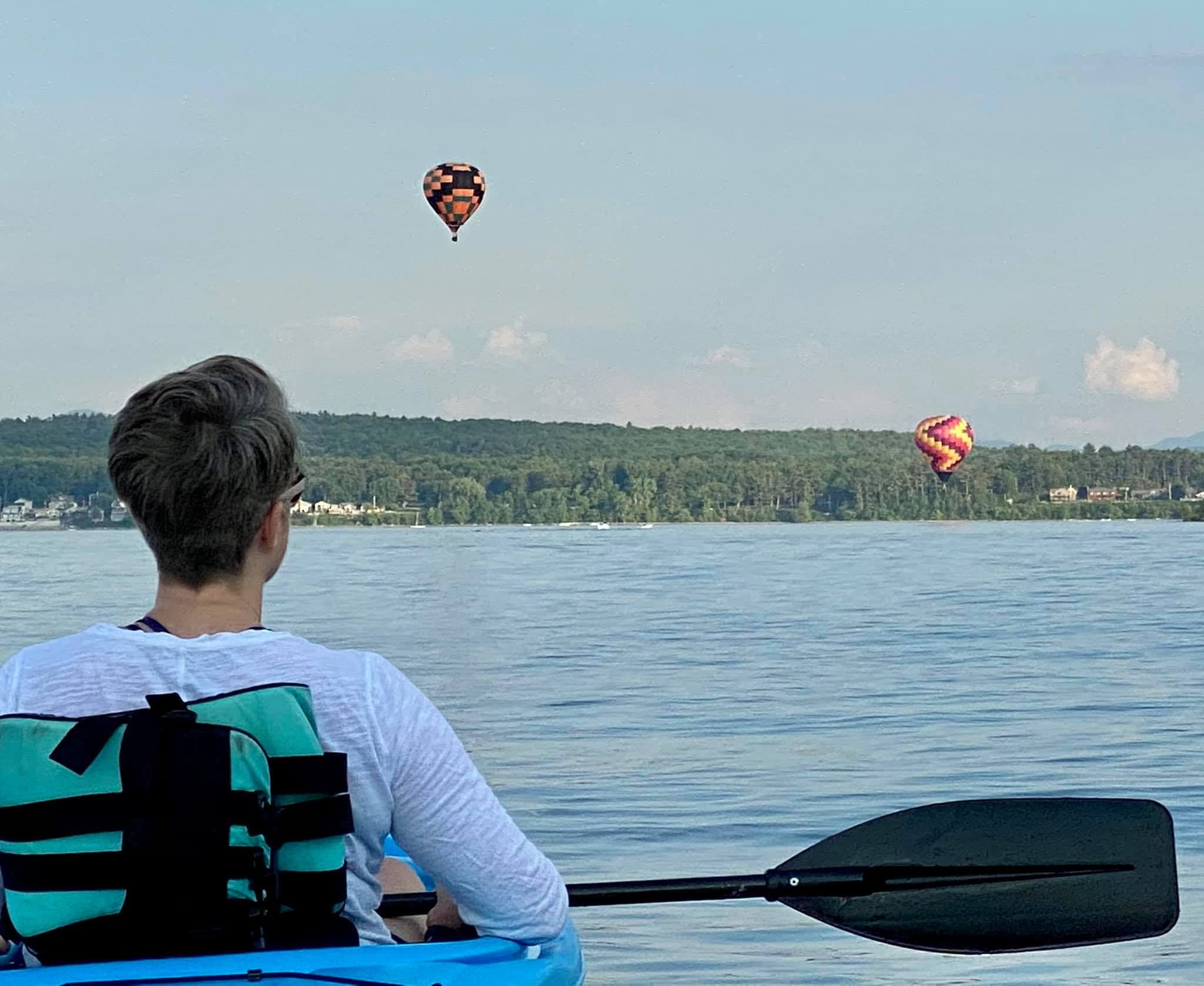 A woman with short gray hair sits in a kayak on a lake with her back to the camera, watching two hot-air balloons fly over the lake.