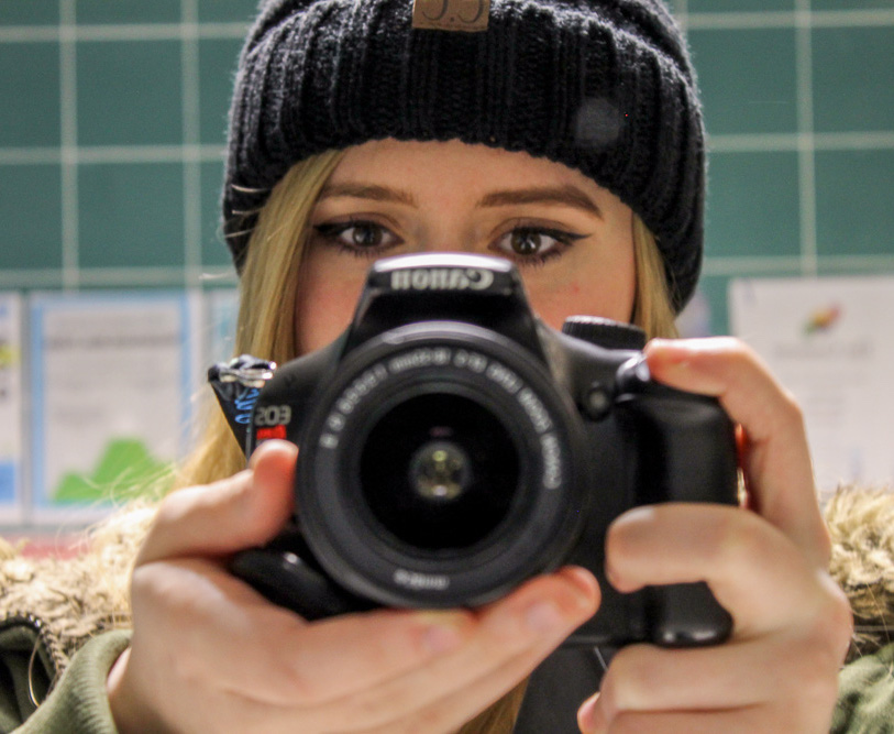 A blond woman with fair skin wearing a knit hat, with the lower half of her face hidden by an SLR camera she holds in her hands
