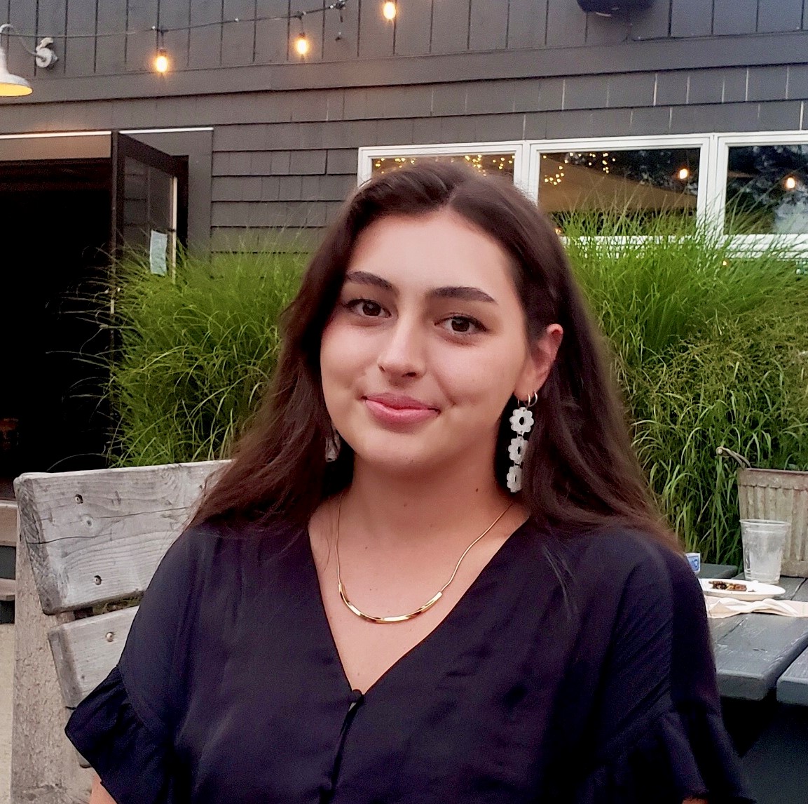 A young woman with fair skin and long dark hair, wearing a black top and long earrings, in front of a house