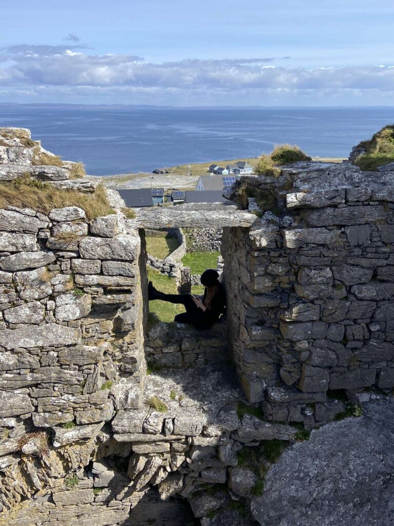 Silhouette of a woman looking at a device while sitting braced in a ruined stone doorway. A coastline with houses is visible in the background.