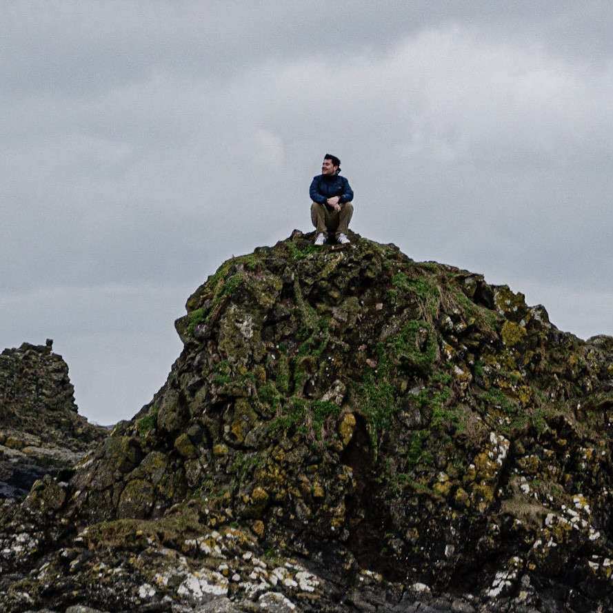 A young man with fair skin and dark hair sitting on top of a rocky outcrop against a cloudy sky