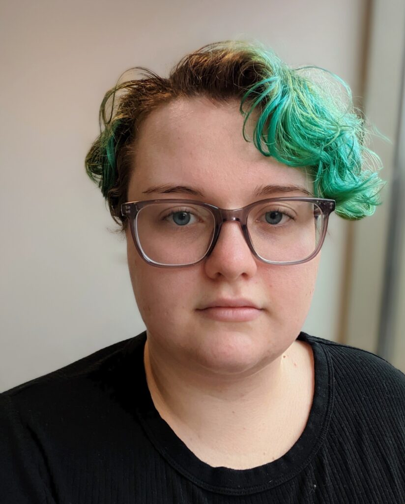 A person with fair skin, short wavy brown and teal hair, and brown glasses, wearing a black shirt