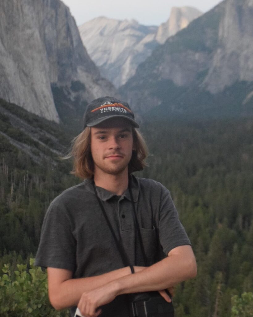 A person with fair skin, shoulder length light brown hair, and a light beard wearing a baseball cap and polo shirt standing in front of a vista of trees and mountains