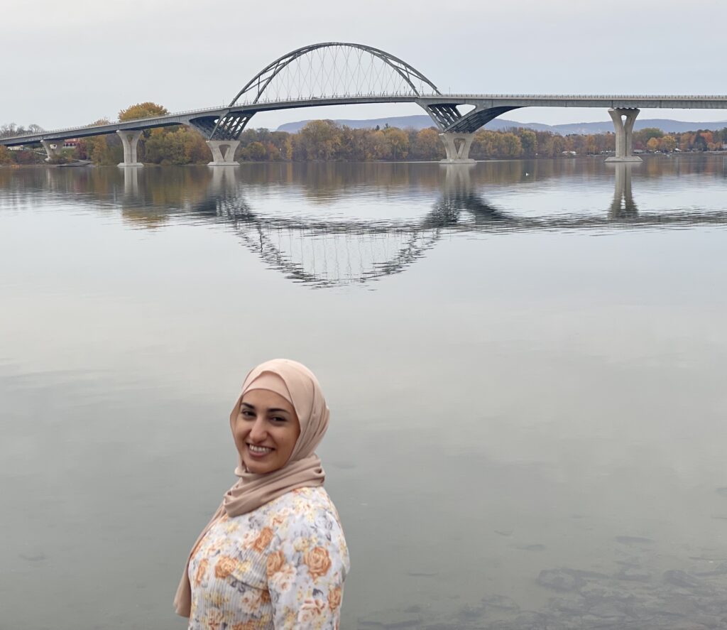 A woman with light skin and dark brows, wearing a flowered top and a beige headscarf, stands in front of a river with an arched bridge in the background