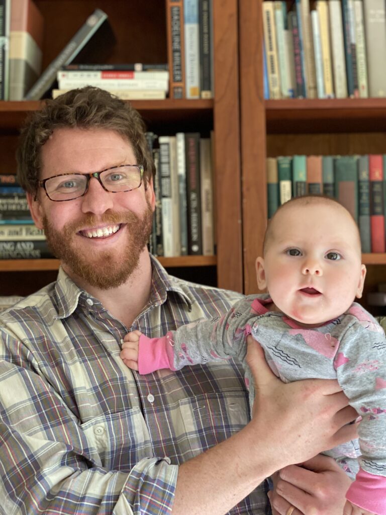 A main with fair skin, reddish hair and beard, and glasses holds a baby while standing in front of bookshelves