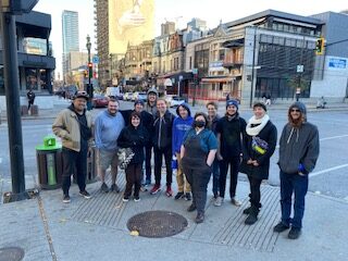 A group of students and faculty in jackets and scarves stand on a street corner in a city, with a mural of Leonard Cohen visible behind them