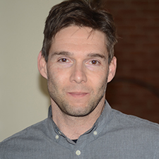 A man with fair skin and dark hair wearing a gray button-down shirt