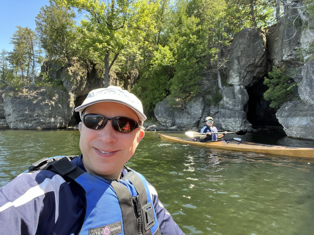 Selfie of a clean-shaven, light-skinned man wearing a baseball cap, sunglasses, and life jacket in a kayak on the water, with another kayaker in the background