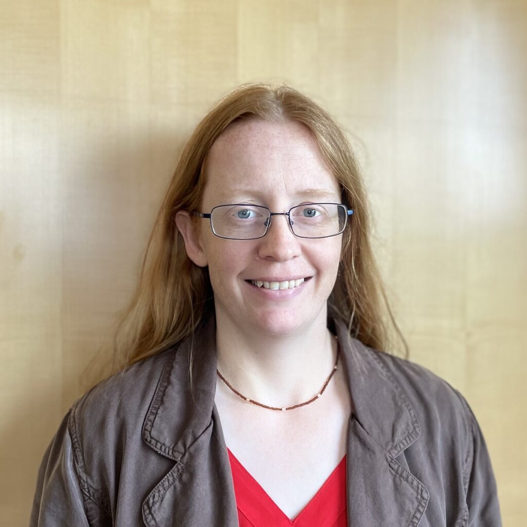 A woman with fair skin and long red hair wearing glasses, a red shirt, and a brown jacket