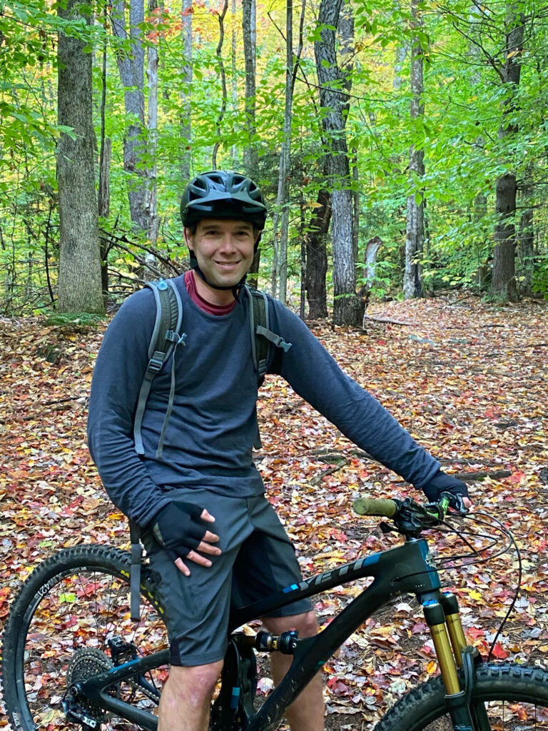 A clean-shaven man with light skin, wearing outdoor clothing, posing on a mountain bike in the forest