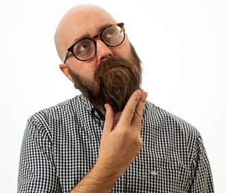 A fair-skinned man with a shaved head, dark beard, and glasses looks up to the side with a thoughtful expression