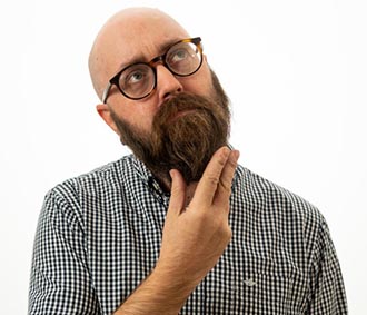 A bald, fair-skinned man with a heavy brown bear and glasses, with his chin in his hand, looking up in a thoughtful manner